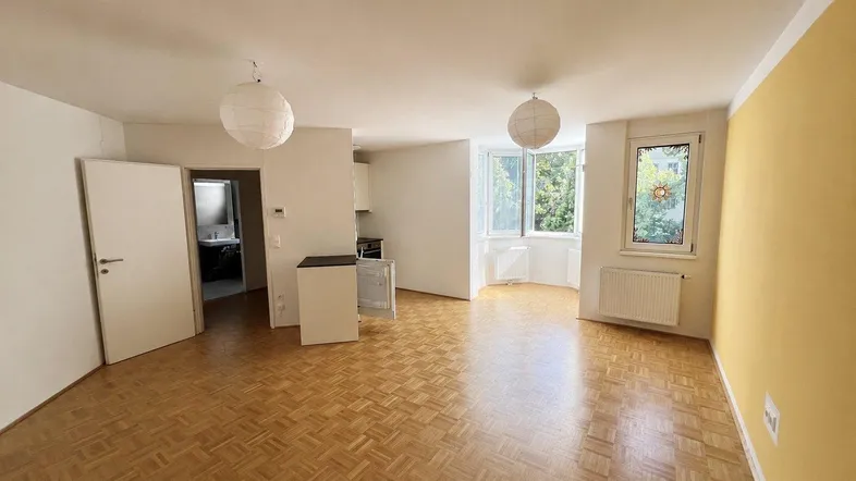 Helles Wohnzimmer mit Parkettboden, gelber Akzentwand und Blick ins Grüne durch die Fenster.