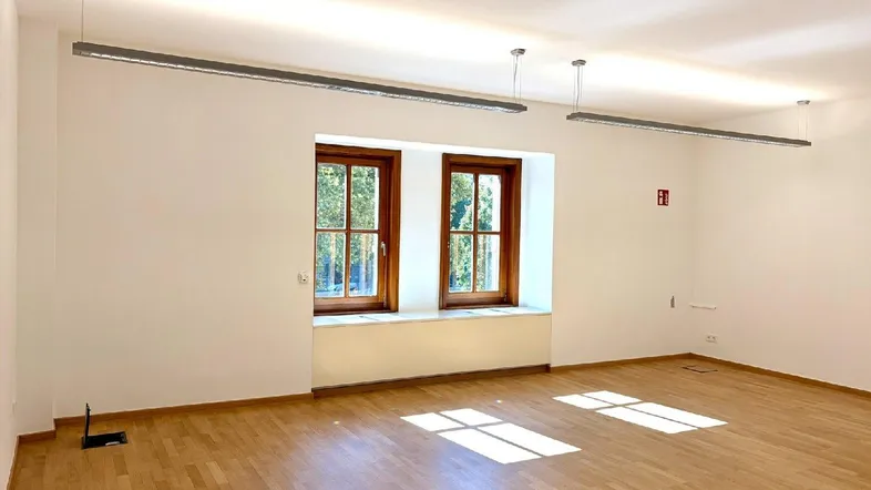 Heller, leerer Raum mit Holzboden, weißen Wänden und zwei Fenstern, durch die Sonnenlicht fällt.