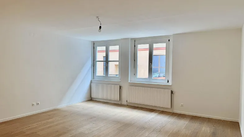 Helles, leeres Zimmer mit Parkettboden und zwei Fenstern, bereit zur individuellen Gestaltung.