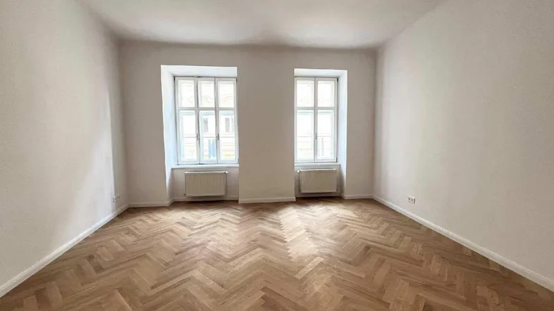 Helles, leeres Zimmer mit Fischgrätparkett und zwei Fenstern, bereit zur individuellen Gestaltung.