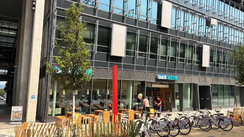 Modernes Geschäftsgebäude mit Glasfassade und Außenbereich, davor Fahrräder und eine kleine Bepflanzung.