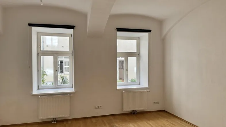 Helles Zimmer mit Holzboden und zwei Fenstern, die auf einen Innenhof blicken.
