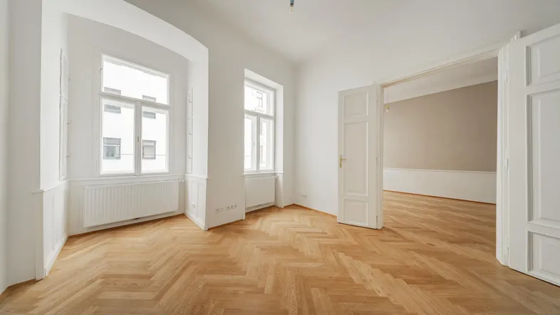 Helles, leeres Zimmer mit Fischgrätparkett und hohen Fenstern, bereit zum Einzug.