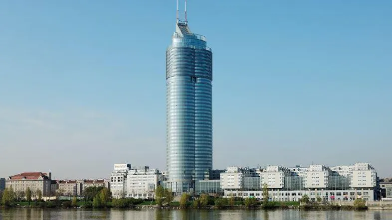 Beeindruckender Wolkenkratzer mit Glasfassade am Flussufer, umgeben von weiteren Gebäuden und Grünflächen.