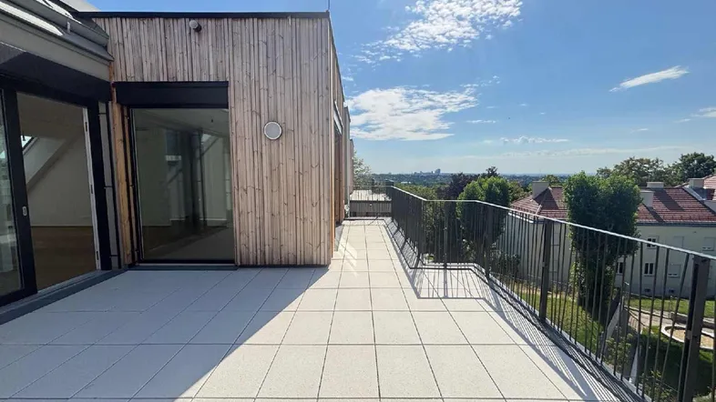Geräumige Dachterrasse mit hellen Fliesen, Holzfassade und weitem Blick ins Grüne unter blauem Himmel.