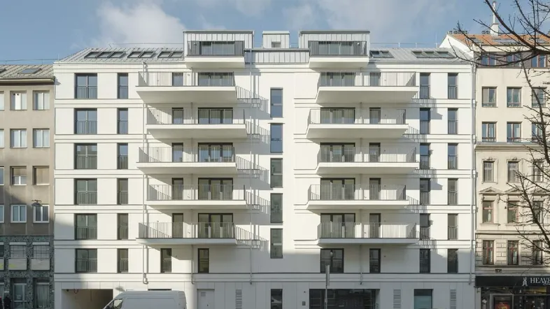 Moderne weiße Fassade eines Neubaus mit zahlreichen Balkonen und großen Fenstern in urbaner Umgebung.