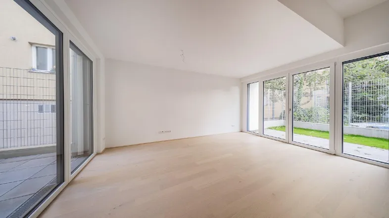 Helles, leeres Zimmer mit großen Fenstern und Zugang zu einer Terrasse und einem Garten.