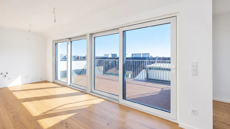 Helles, leeres Zimmer mit Holzboden und großen Schiebetüren, die auf eine Dachterrasse führen.