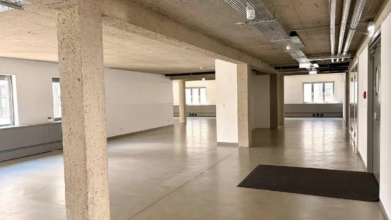 Großer, offener Büroraum mit Sichtbetondecke, freiliegenden Leitungen und großen Fenstern.