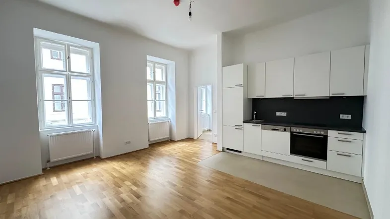 Heller Wohnbereich mit integrierter Küchenzeile, Parkettboden und großen Fenstern, die viel Licht hereinlassen.