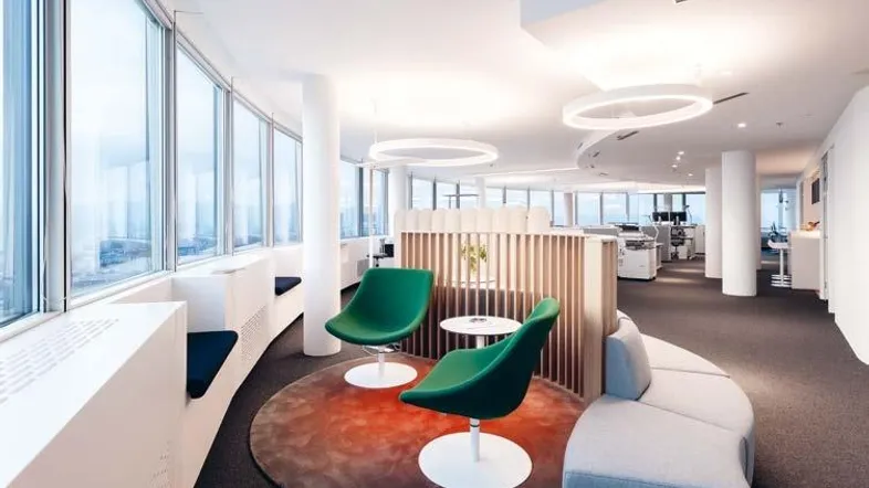 Modernes Büro mit großen Fenstern, grünen Sesseln und einem runden Teppich als gemütliche Lounge-Ecke.
