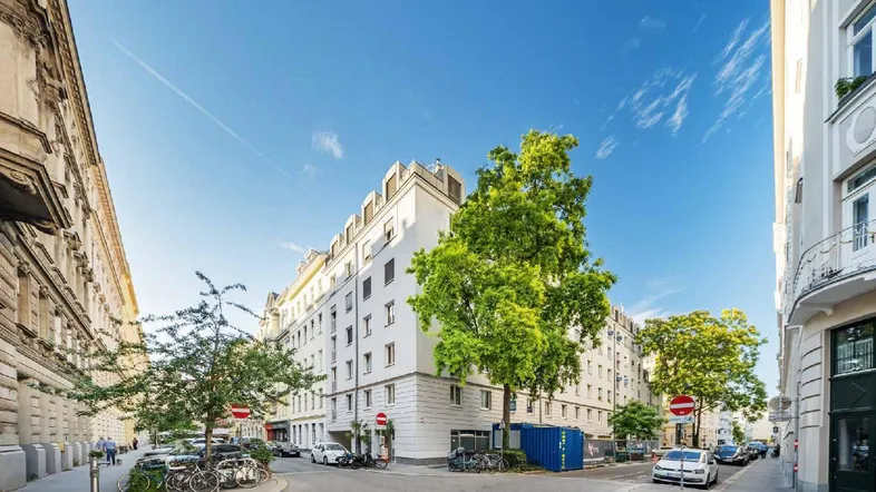 Weißes Gebäude an einer Straßenecke mit Bäumen und blauem Himmel in urbaner Umgebung.