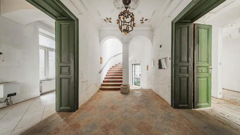 Historischer Eingangsbereich mit hohen Decken, Stuckverzierungen und grünen Flügeltüren.
