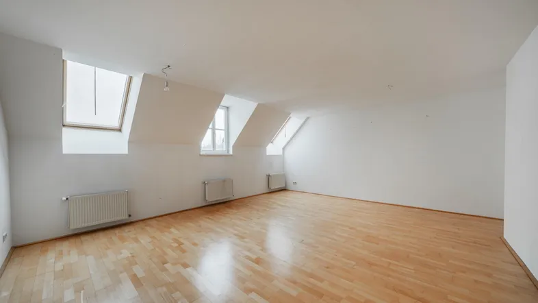 Helles Zimmer mit Dachschrägen, Holzfußboden und mehreren Fenstern, ideal als Wohn- oder Schlafbereich.
