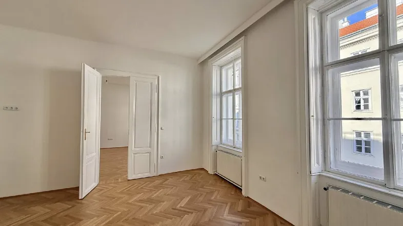 Helles Zimmer mit Fischgrätparkettboden, hohen Fenstern und weiß gestrichenen Wänden, bereit zum Einzug.