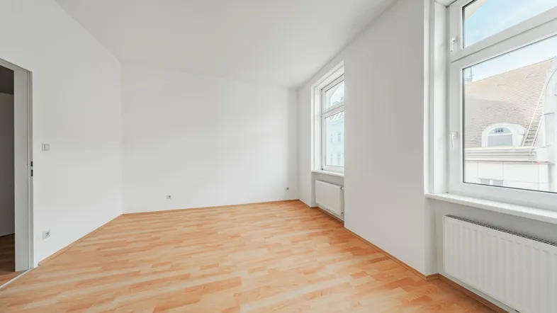 Helles, leeres Zimmer mit Laminatboden und zwei Fenstern, bereit zur individuellen Gestaltung.