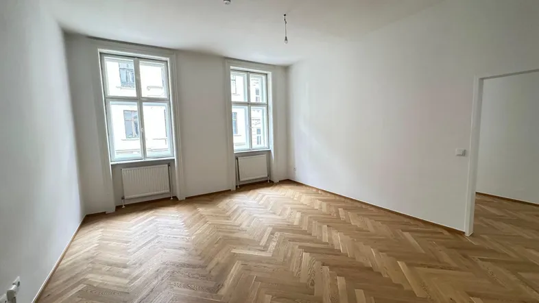 Helles Zimmer mit Fischgrätparkett, weißen Wänden und zwei großen Fenstern.