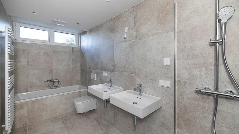 Modernes Badezimmer mit zwei Waschbecken, Badewanne, Dusche, WC und Fenster, hell gefliest.