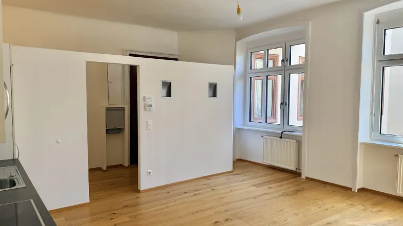 Helles Zimmer mit Holzboden, großen Fenstern und einem abgetrennten Bereich für eine Küchenzeile.