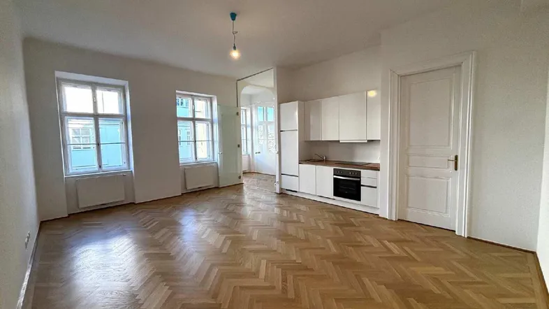 Helles Wohnzimmer mit Fischgrätparkett, zwei Fenstern und integrierter weißer Küchenzeile.