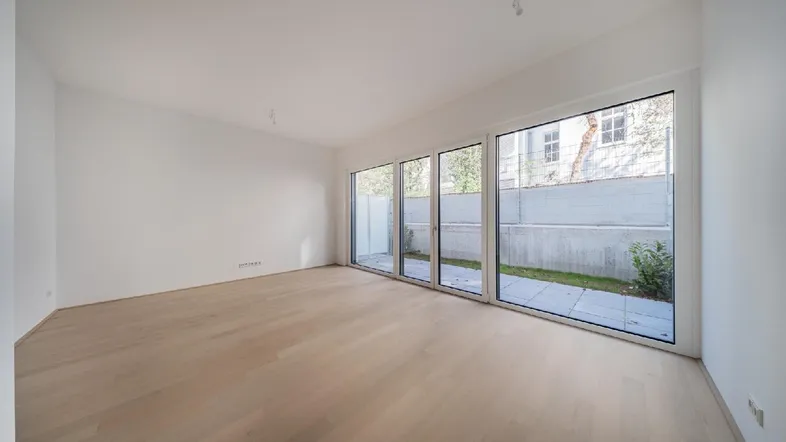 Helles, leeres Zimmer mit großen Fenstern und Zugang zu einem kleinen Gartenbereich.