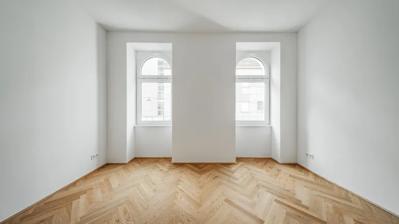 Helles Zimmer mit Fischgrätparkett und zwei Rundbogenfenstern, bereit zur individuellen Gestaltung.