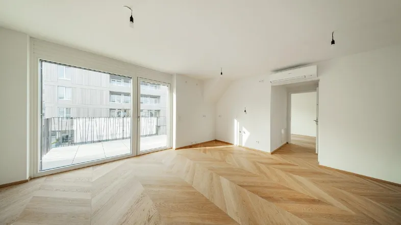 Helles, leeres Zimmer mit modernem Fischgrätparkett und Zugang zu einem Balkon.