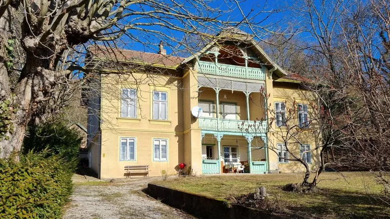 Historische Villa mit gelber Fassade und grünem Balkon, umgeben von Bäumen unter blauem Himmel.