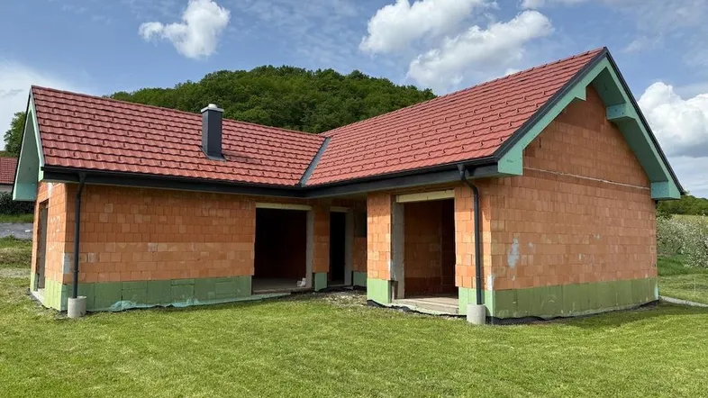 Neubau eines Einfamilienhauses im Rohbau mit rotem Ziegeldach und grüner Isolierung, umgeben von Natur.