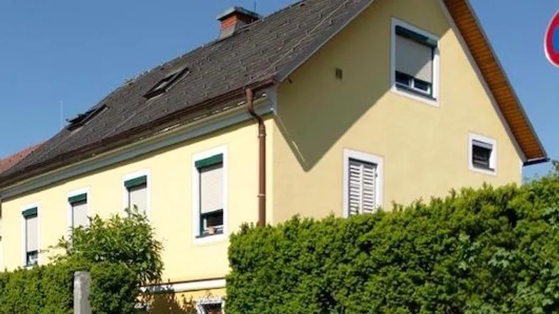 Seitenansicht des gelben Hauses mit grüner Hecke und Rollläden an den Fenstern bei Sonnenschein.