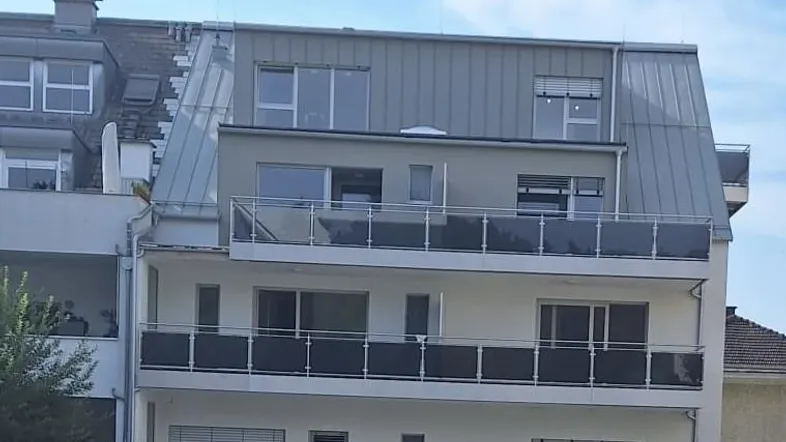 Modernes Mehrfamilienhaus mit mehreren Balkonen und einem Dachgeschossausbau unter blauem Himmel.