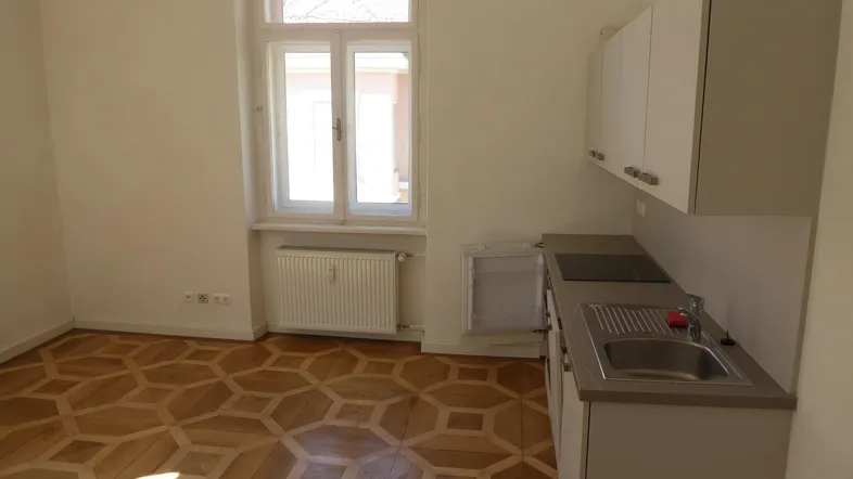 Helle Küchenzeile mit integriertem Kühlschrank, Kochfeld und Spüle, dazu ein Fenster mit Tageslicht.
