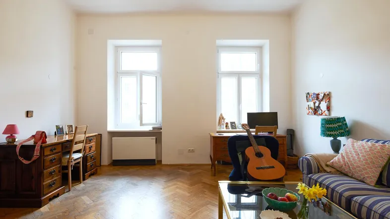 Helles Wohnzimmer mit Parkettboden, zwei Fenstern und gemütlicher Sitzecke.