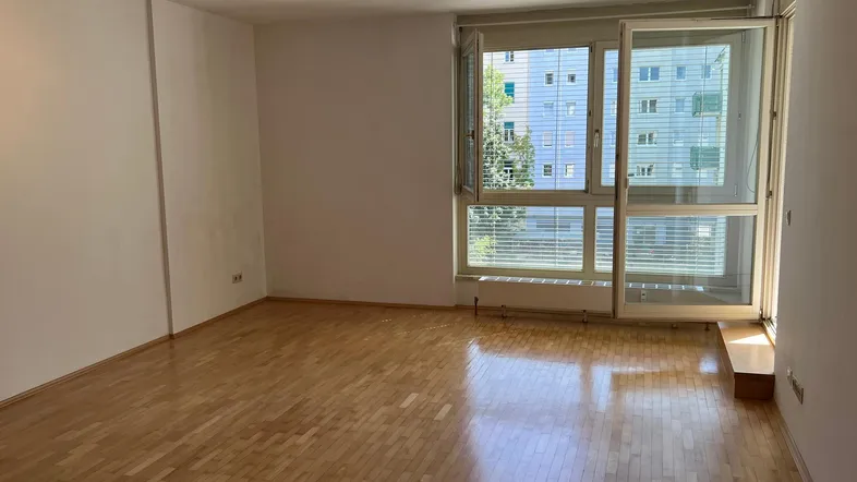 Helles, leeres Wohnzimmer mit Parkettboden und großen Fenstern, die viel Tageslicht hereinlassen.
