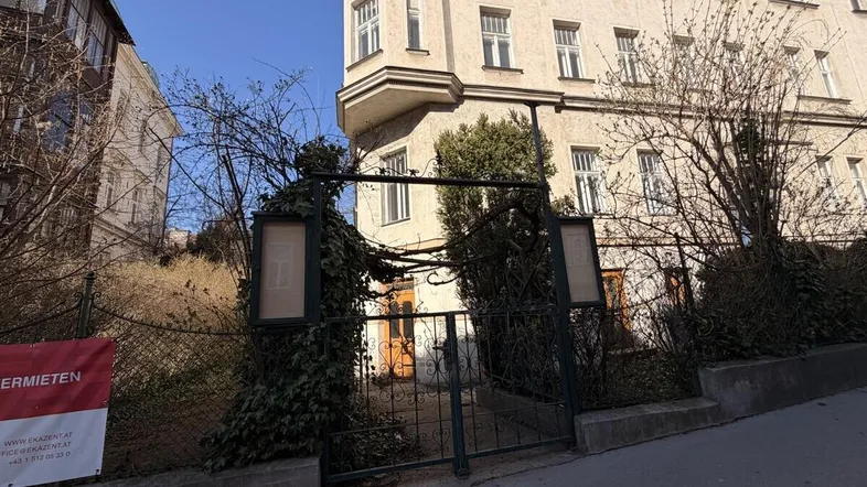 Gebäudeansicht mit historischer Fassade und Eingangsbereich, umgeben von kahlen Bäumen und einem Zaun an einem sonnigen Tag.