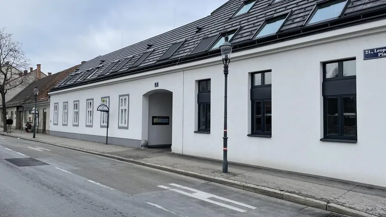 Langes, weißes Gebäude mit modernem Dach und vielen Dachfenstern an einer ruhigen Straße.