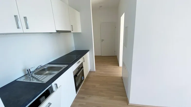 Moderne, kompakte Küche mit weißen Schränken, dunkler Arbeitsplatte und integriertem Backofen, offen zum Flur.