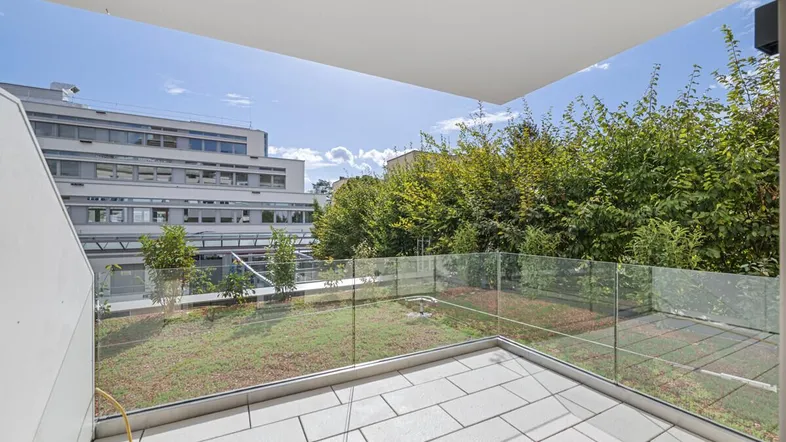 Geräumige Terrasse mit modernen Fliesen, Glasgeländer und Blick auf eine begrünte Fläche.