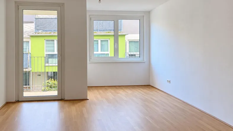 Helles, leeres Zimmer mit Parkettboden und Zugang zu einem Balkon, mit Blick auf grüne Fassaden.