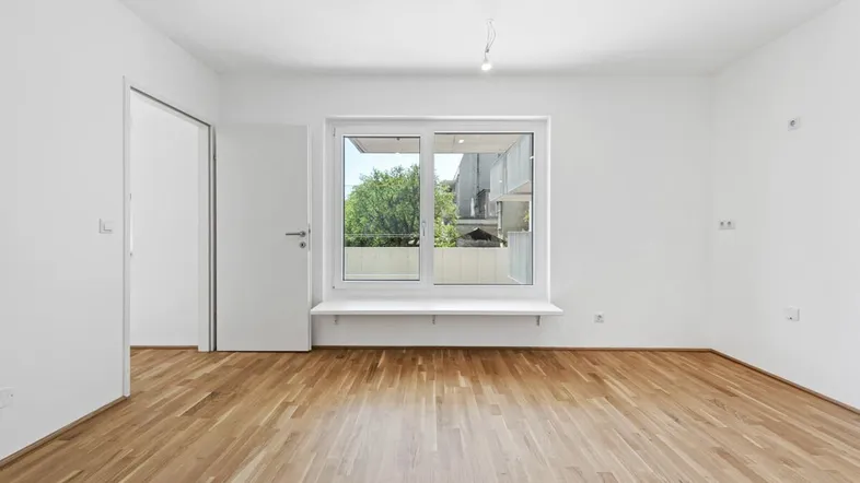 Helles, leeres Zimmer mit Parkettboden und großem Fenster mit Blick ins Grüne.