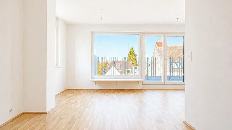 Helles, leeres Zimmer mit Parkettboden und großem Fenster, das Zugang zu einem Balkon bietet.