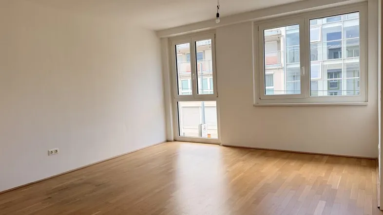 Helles Zimmer mit Holzboden und großen Fenstern, die viel Tageslicht hereinlassen.