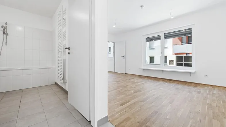 Blick von einem leeren Raum mit Parkettboden in ein Badezimmer mit Dusche und Handtuchheizkörper.