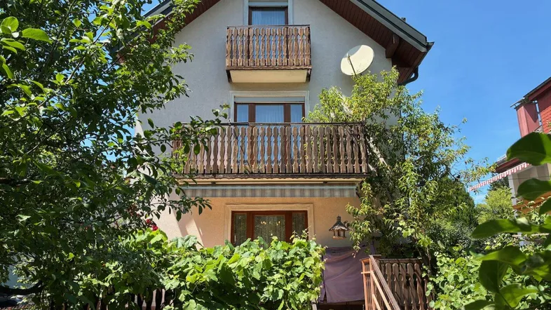 Vorderansicht des Hauses mit zwei Holzbalkonen und üppiger Vegetation im Vorgarten.