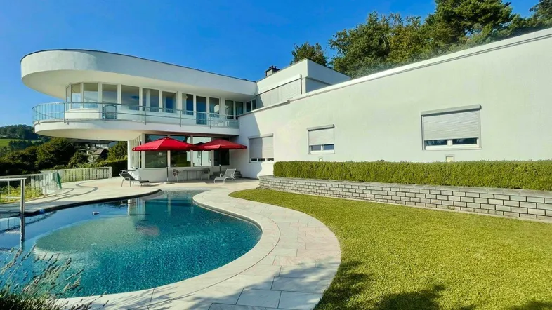 Moderne Villa mit geschwungener Architektur, großem Pool und gepflegter Rasenfläche unter blauem Himmel.