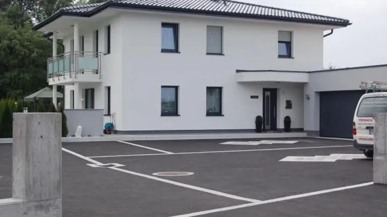 Modernes weißes Einfamilienhaus mit dunklem Dach, Garage und gepflasterter Einfahrt mit Parkplätzen.