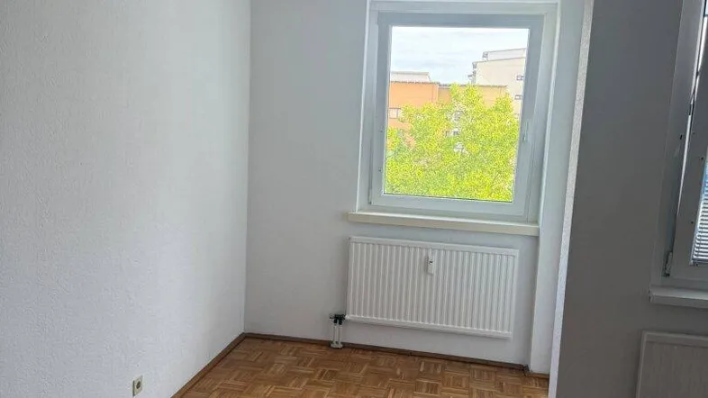 Helles Zimmer mit Parkettboden, weißen Wänden und Blick ins Grüne durch das Fenster.