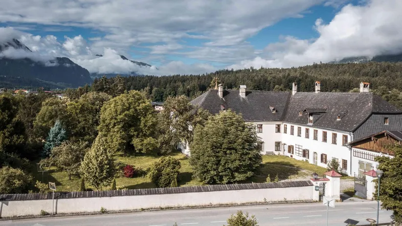 Historisches Gebäude mit traditioneller Architektur, umgeben von Bäumen und einer Mauer, mit Bergen im Hintergrund.
