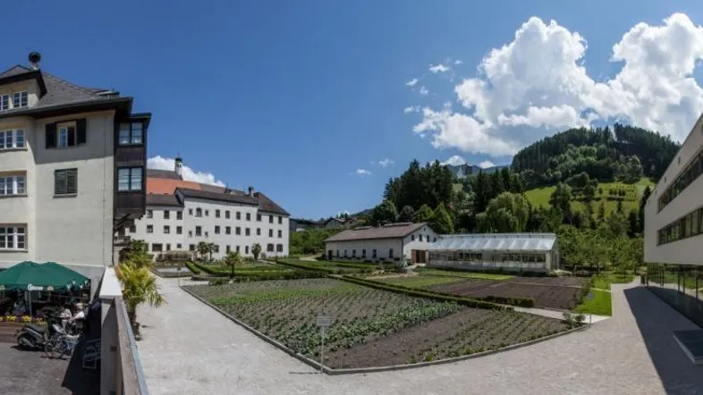 Panoramablick auf ein Anwesen mit traditionellen und modernen Gebäuden, einem gepflegten Garten und Bergen im Hintergrund.
