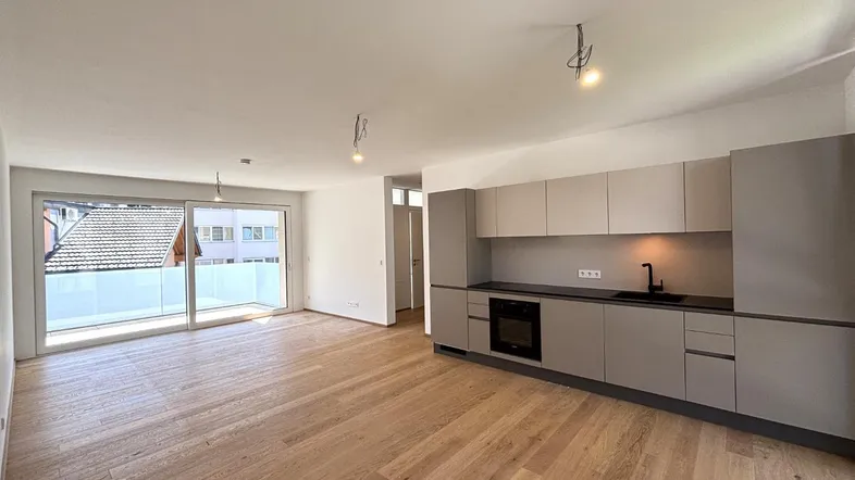 Heller offener Wohnbereich mit integrierter Küche, Holzboden und Zugang zu einem Balkon.